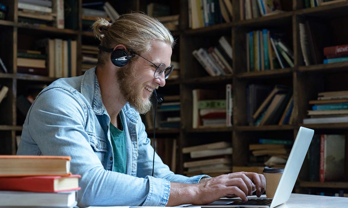 Ein Mann mit blonden nach hinten gebundenen Haaren sitzt in einem Raum mit Büchern im Hintergrund und tippt an seinem Laptop.