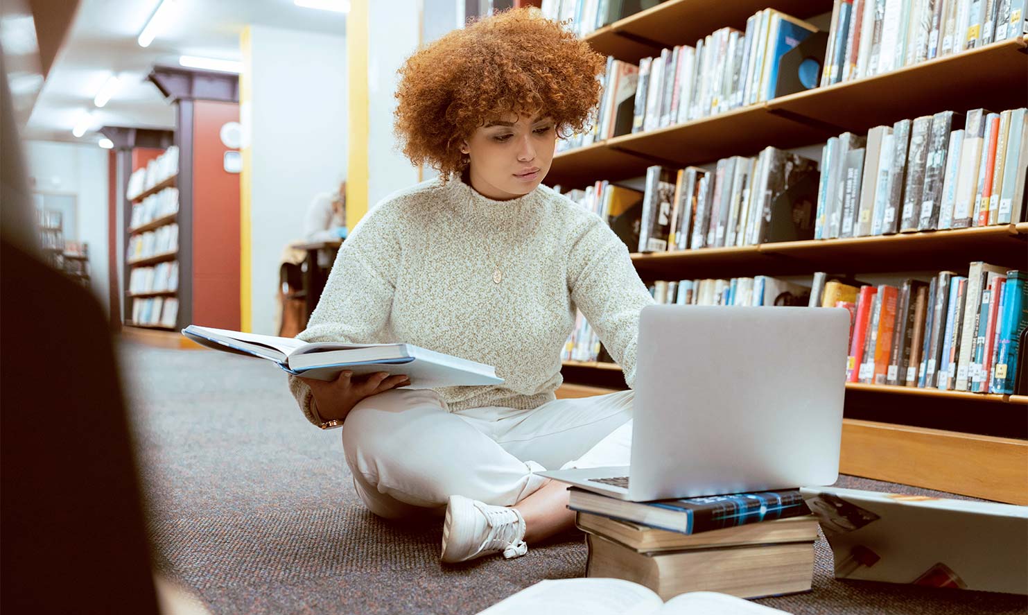 Eine junge Frau mit lockigen Haaren sitzt in einer Bibliothek auf dem Boden während sie ihren Laptop aufgeklappt auf einem Stapel Bücher hat.