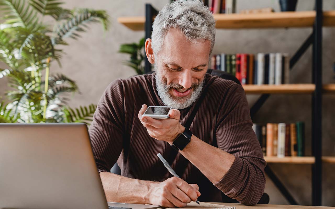 Ein grauhaariger Mann mit Bart sitzt an seinem Arbeitsplatz am Laptop und telefoniert mit seinem Handy mit Lautsprecher.